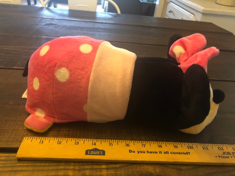 May include: A pink and black plush toy shaped like a cartoon mouse. The mouse is lying on its side with its head resting on the table. The toy has white polka dots on its pink body and a pink bow on its head.