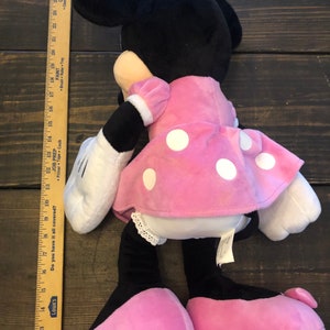 May include: A pink and black plush toy of Minnie Mouse. She is wearing a pink dress with white polka dots and a white bow. She has a black nose and big black eyes.