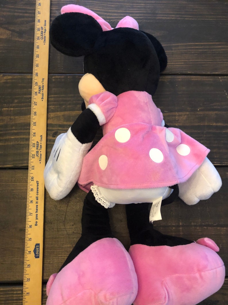 May include: A pink and black plush toy of Minnie Mouse. She is wearing a pink dress with white polka dots and a white bow. She has a black nose and big black eyes.