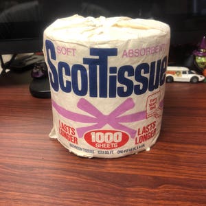 May include: A white roll of ScotTissue bathroom tissue with a purple ribbon and the text "Soft Absorbent ScotTissue" in blue. The roll has 1000 sheets and is one-ply, 4.5 inches by 4.4 inches.