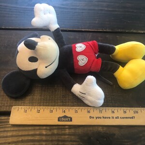 Puede incluir: Un juguete de peluche Mickey Mouse negro y rojo con una cara blanca y zapatos amarillos. El juguete est&aacute; acostado sobre una superficie de madera con una regla debajo. La regla tiene las palabras "Do you have it all covered?" y "LOWE'S" impresas en ella.