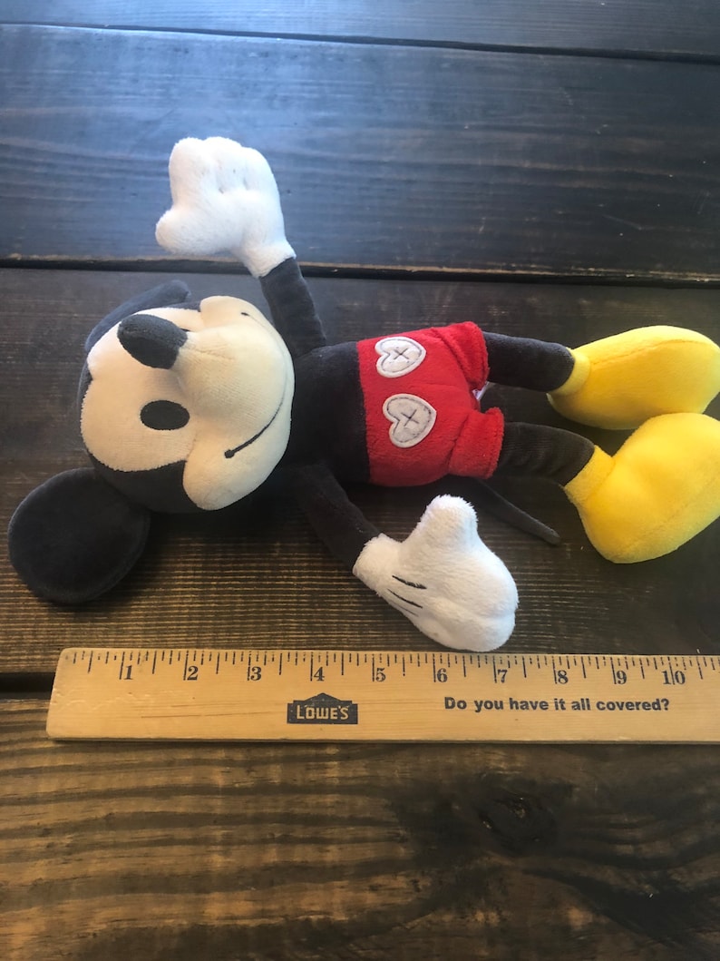 Puede incluir: Un juguete de peluche Mickey Mouse negro y rojo con una cara blanca y zapatos amarillos. El juguete est&aacute; acostado sobre una superficie de madera con una regla debajo. La regla tiene las palabras "Do you have it all covered?" y "LOWE'S" impresas en ella.