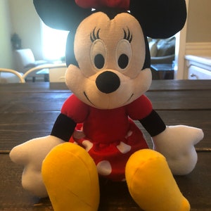 May include: A plush toy of Minnie Mouse, wearing a red dress with white polka dots and a red bow. She has black ears and a black nose, and her eyes are closed.