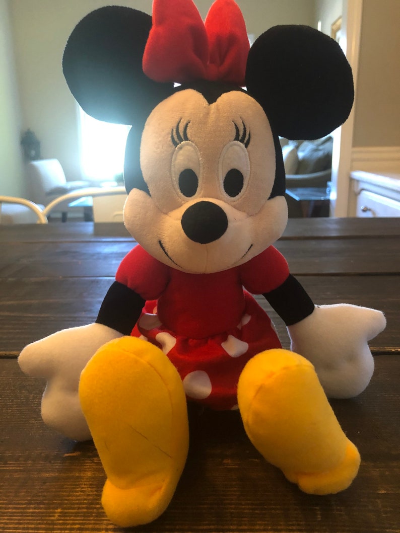 May include: A plush toy of Minnie Mouse, wearing a red dress with white polka dots and a red bow. She has black ears and a black nose, and her eyes are closed.