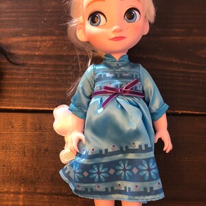 May include: A small doll with blonde hair wearing a blue dress with a purple sash and a snowflake pattern. The doll has blue eyes and is holding a small white stuffed animal.