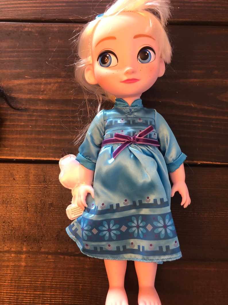 May include: A small doll with blonde hair wearing a blue dress with a purple sash and a snowflake pattern. The doll has blue eyes and is holding a small white stuffed animal.
