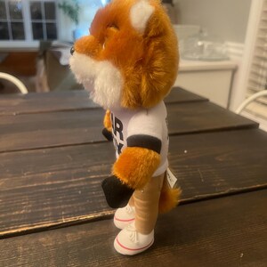 May include: A plush fox toy with orange and white fur, wearing a white t-shirt with black lettering. The fox has tan legs, black paws, and white trainers with red trim. The toy is standing on a dark wooden surface.