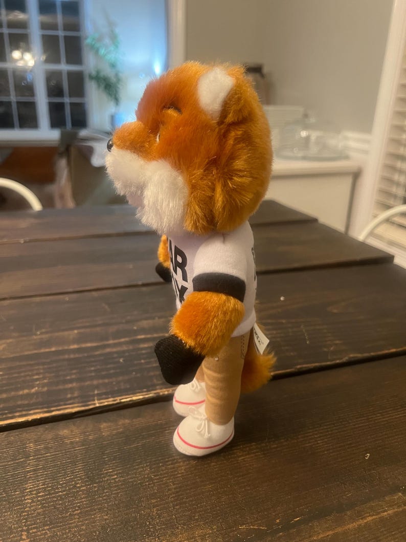 May include: A plush fox toy with orange and white fur, wearing a white t-shirt with black lettering. The fox has tan legs, black paws, and white trainers with red trim. The toy is standing on a dark wooden surface.