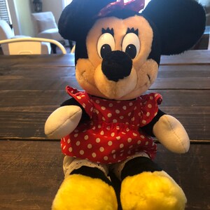 K&ouml;nnte beinhalten: Ein Pl&uuml;schtier von Minnie Mouse, die ein rotes Kleid mit wei&szlig;en Punkten und eine rote Schleife tr&auml;gt. Sie hat schwarze Ohren und eine schwarze Nase. Ihre Augen sind gro&szlig; und schwarz.