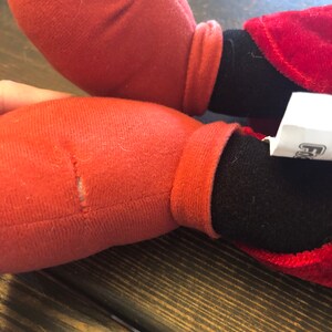 May include: Red and black plush toy with a torn seam on one of the red legs. The toy is sitting on a wooden surface.