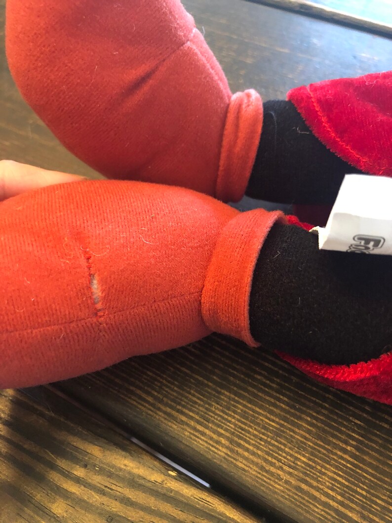May include: Red and black plush toy with a torn seam on one of the red legs. The toy is sitting on a wooden surface.