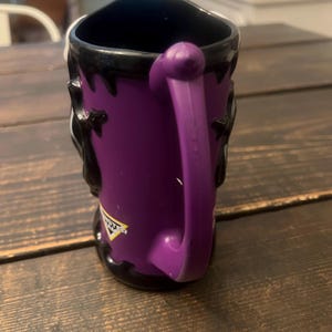 May include: Purple ceramic mug with a black interior and handle. The mug features a design with black accents and a logo on the side. The handle is a matching purple color.