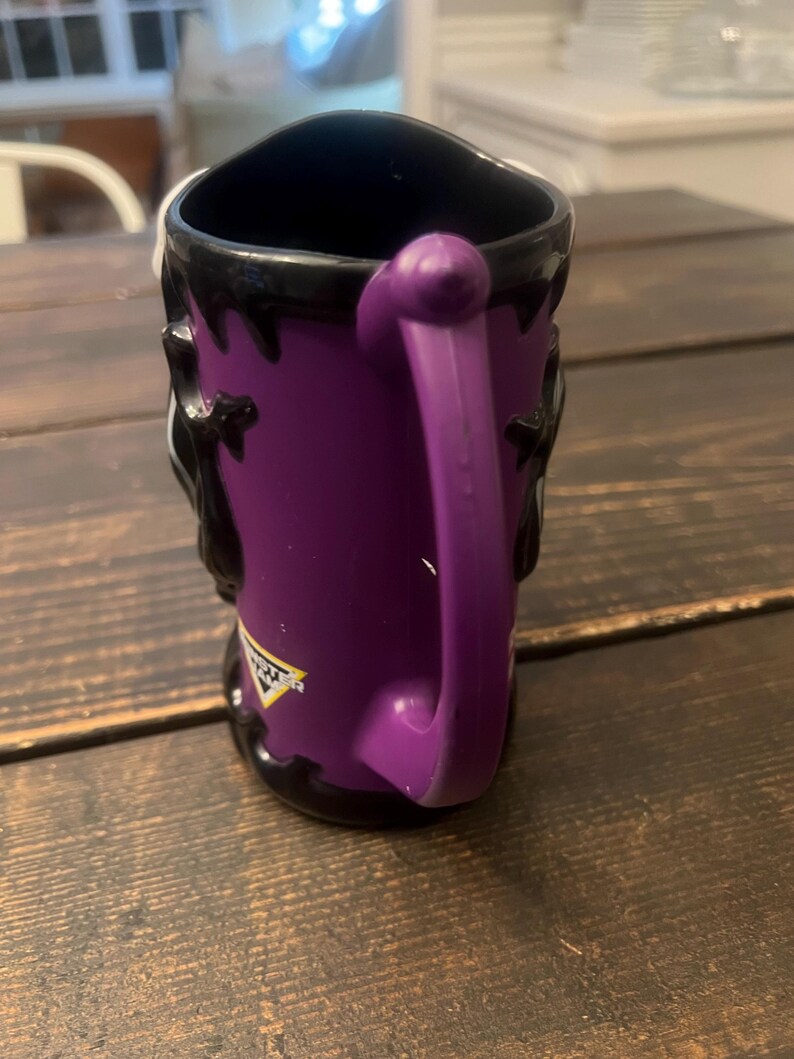 May include: Purple ceramic mug with a black interior and handle. The mug features a design with black accents and a logo on the side. The handle is a matching purple color.