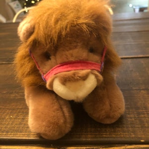 May include: A brown plush lion with a pink and purple collar. The lion is lying on a wooden surface.