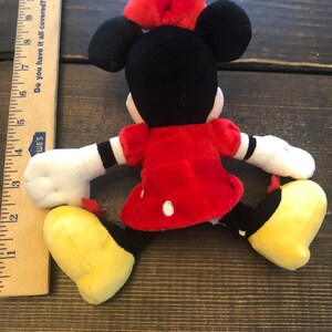 Op de afbeelding: Een rood en zwart pluche speelgoed van een cartoonmuis met een rode strik. De muis draagt een rode jurk met witte stippen en heeft gele schoenen.