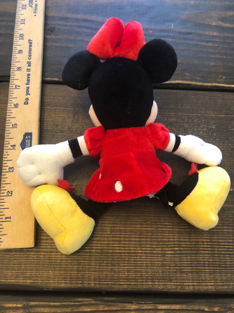 Op de afbeelding: Een rood en zwart pluche speelgoed van een cartoonmuis met een rode strik. De muis draagt een rode jurk met witte stippen en heeft gele schoenen.