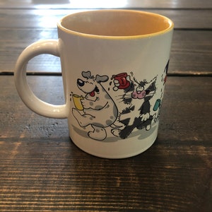 May include: A white ceramic coffee mug with a yellow rim. The mug features a cartoon illustration of a dog and a cat holding coffee cups. The dog is gray with black spots and the cat is black and white. The text on the mug reads 