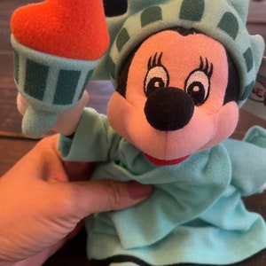 May include: A plush toy of Mickey Mouse dressed as the Statue of Liberty. The toy is holding a miniature replica of the Statue of Liberty's torch in its right hand. The toy is wearing a green dress with a crown and a red mouth.