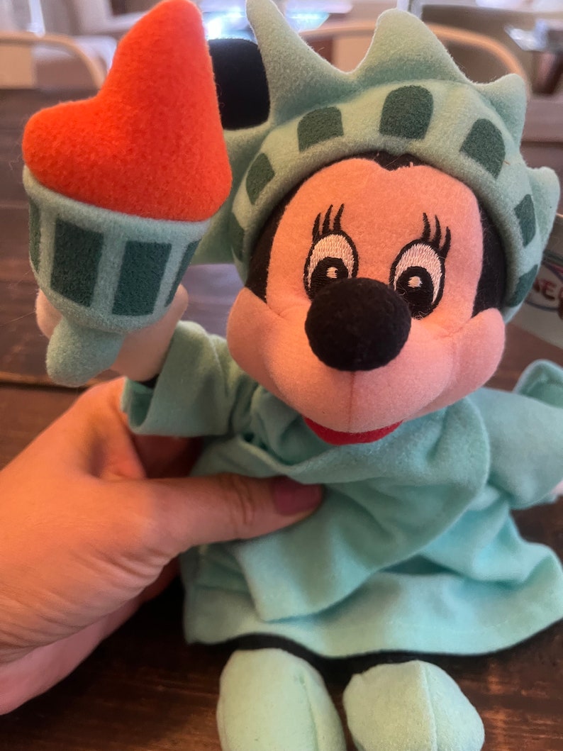 May include: A plush toy of Mickey Mouse dressed as the Statue of Liberty. The toy is holding a miniature replica of the Statue of Liberty's torch in its right hand. The toy is wearing a green dress with a crown and a red mouth.