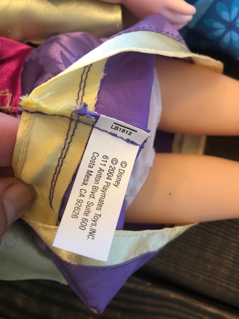 May include: A purple and yellow doll dress with a white label that reads "&copy; Disney &copy;2004 Playmates Toys, INC. 611 Anton Blvd. Suite 600 Costa Mesa, CA 92626" and "LB1813".