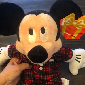 May include: A plush Mickey Mouse toy wearing red and green plaid pajamas. The toy has a black nose, large black ears, and white eyes. The toy has a Disney Store tag attached to its right ear.