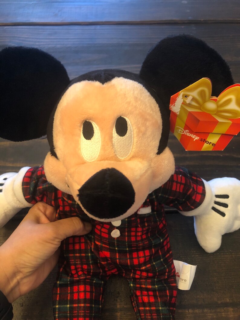 May include: A plush Mickey Mouse toy wearing red and green plaid pajamas. The toy has a black nose, large black ears, and white eyes. The toy has a Disney Store tag attached to its right ear.