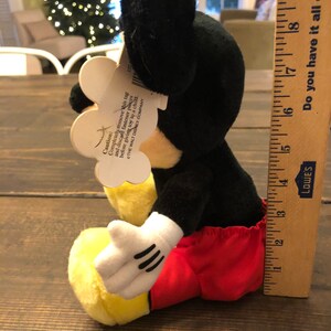 May include: A black and yellow plush Mickey Mouse toy wearing red shorts. The toy is sitting on a wooden surface. The tag attached to the toy reads "Caution: Completely remove this tag and discard fastener pieces before giving toy to a child. THE WALT DISNEY COMPANY."