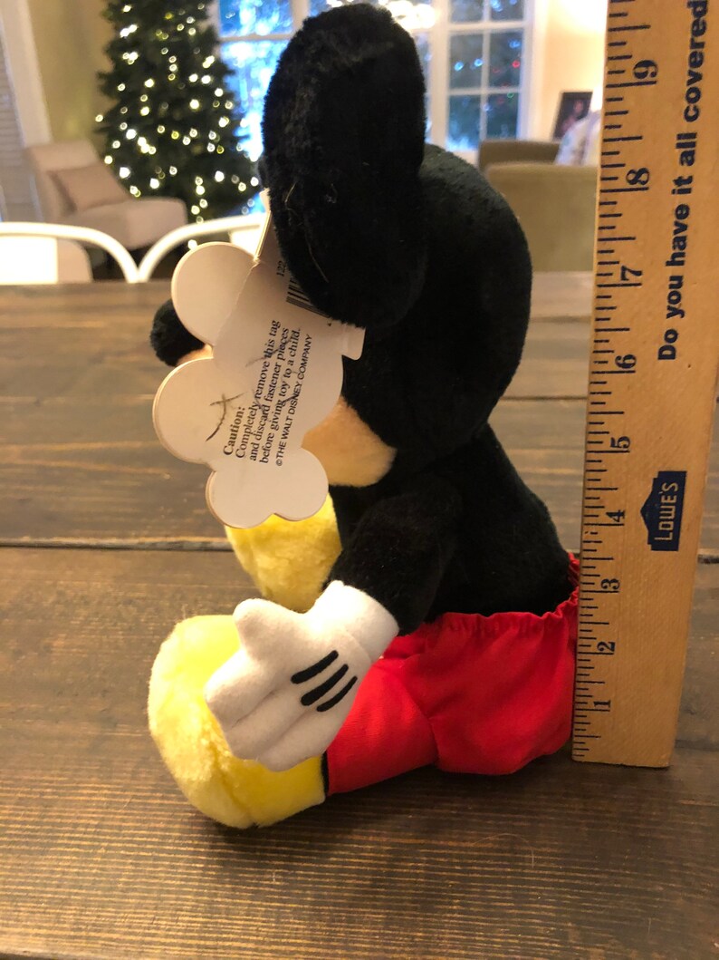May include: A black and yellow plush Mickey Mouse toy wearing red shorts. The toy is sitting on a wooden surface. The tag attached to the toy reads "Caution: Completely remove this tag and discard fastener pieces before giving toy to a child. THE WALT DISNEY COMPANY."