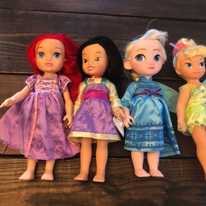 May include: Four Disney princess dolls, including Ariel, Mulan, Elsa, and Tinkerbell. The dolls are wearing their signature dresses and have detailed facial features.