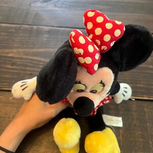 May include: A black and white plush toy of Minnie Mouse with a red and white polka dot bow. The toy is sitting on a wooden surface.