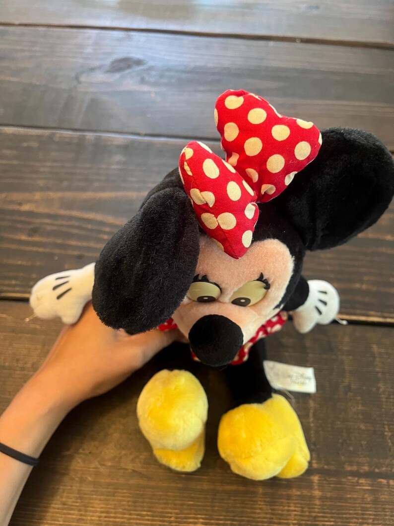 May include: A black and white plush toy of Minnie Mouse with a red and white polka dot bow. The toy is sitting on a wooden surface.