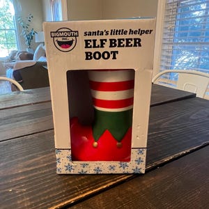 May include: An Elf Beer Boot in a box. The boot is red with a green cuff and gold accents. The upper portion of the boot is white with red stripes. The box reads "santa's little helper" and "ELF BEER BOOT".