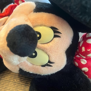 May include: A plush toy of Minnie Mouse with black ears, a red bow with white polka dots, and large yellow eyes with black eyelashes.