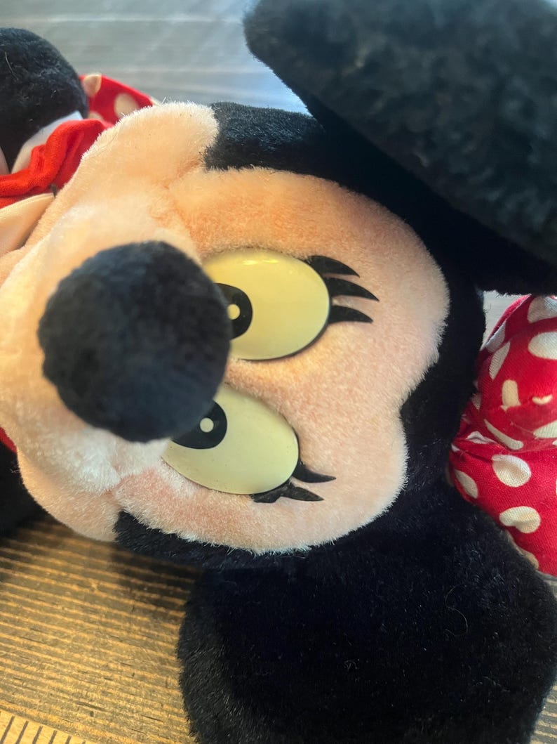 May include: A plush toy of Minnie Mouse with black ears, a red bow with white polka dots, and large yellow eyes with black eyelashes.