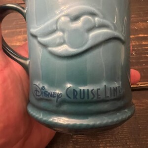 May include: A teal ceramic mug with a handle. The mug features a raised Mickey Mouse design and the words "Disney Cruise Line" in dark blue lettering. The mug has a ridged base.
