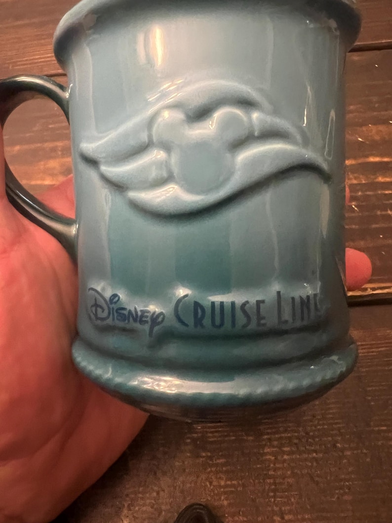 May include: A teal ceramic mug with a handle. The mug features a raised Mickey Mouse design and the words "Disney Cruise Line" in dark blue lettering. The mug has a ridged base.