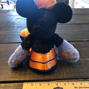 May include: A plush Minnie Mouse doll in a Halloween costume. The doll has black fur, large round ears, and a bow on her head. She wears a purple and orange outfit with star accents. The doll is approximately 10 inches tall.