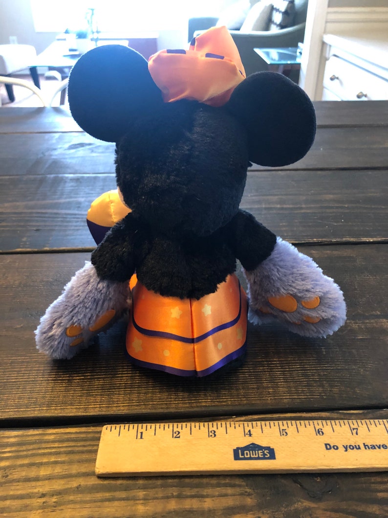 May include: A plush Minnie Mouse doll in a Halloween costume. The doll has black fur, large round ears, and a bow on her head. She wears a purple and orange outfit with star accents. The doll is approximately 10 inches tall.