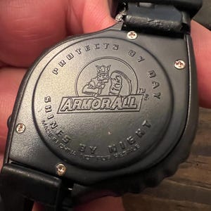 May include: Black watch with the text "PROTECTS BY DAY" and "SHINES BY NIGHT". The watch face features the "ARMOR ALL" logo with a knight graphic. The watch is made in China.