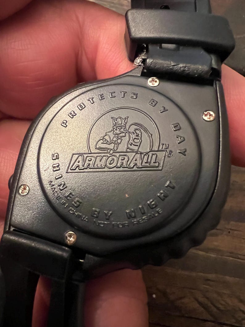 May include: Black watch with the text "PROTECTS BY DAY" and "SHINES BY NIGHT". The watch face features the "ARMOR ALL" logo with a knight graphic. The watch is made in China.