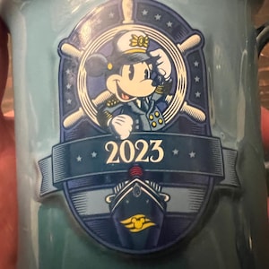 May include: A light blue ceramic mug featuring a nautical-themed design with Minnie Mouse in a captain's uniform. The mug has a ship's wheel and the year "2023" printed on it. The mug has a handle and is likely for beverages.