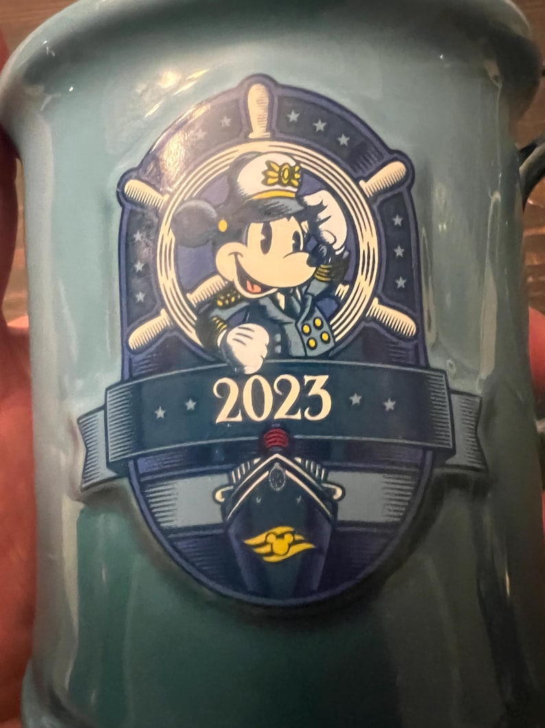 May include: A light blue ceramic mug featuring a nautical-themed design with Minnie Mouse in a captain's uniform. The mug has a ship's wheel and the year "2023" printed on it. The mug has a handle and is likely for beverages.