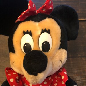 K&ouml;nnte beinhalten: Ein Pl&uuml;schtier von Minnie Mouse, die ein rotes Kleid mit wei&szlig;en Punkten und eine rote Schleife mit wei&szlig;en Punkten tr&auml;gt. Sie hat gro&szlig;e schwarze Augen und eine schwarze Nase.