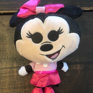 May include: A plush toy of Minnie Mouse wearing a pink dress and a pink bow. She has black eyes, a black nose, and a pink mouth. The toy is about 10 inches tall.