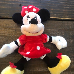 Op de afbeelding: Een pluche speelgoed van Minnie Mouse die een rode jurk draagt met witte stippen en een rode strik met witte stippen. Ze heeft zwarte schoenen met gele zolen en rode strikjes.