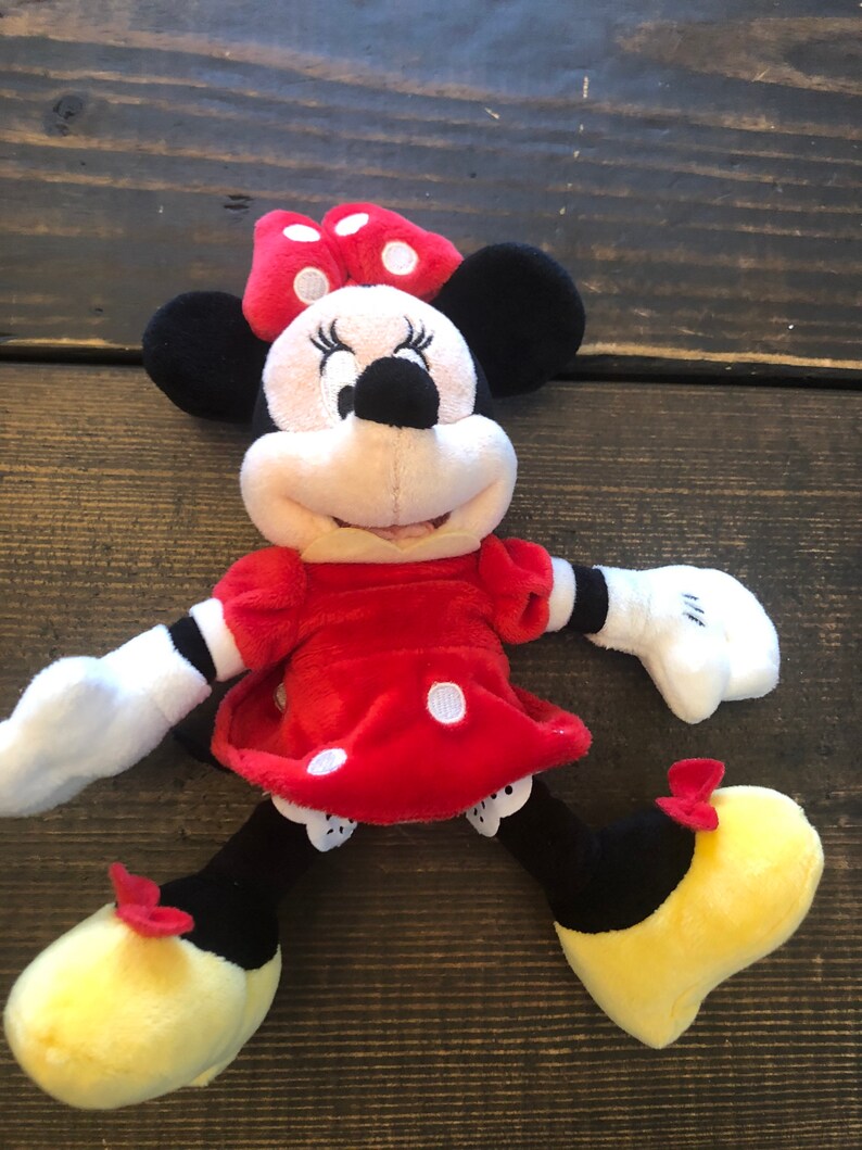 Op de afbeelding: Een pluche speelgoed van Minnie Mouse die een rode jurk draagt met witte stippen en een rode strik met witte stippen. Ze heeft zwarte schoenen met gele zolen en rode strikjes.