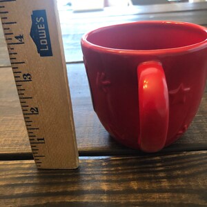 May include: A red ceramic mug with a star design and a handle. The mug is sitting on a wooden surface.