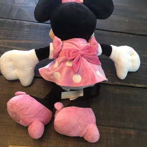 May include: A pink and black plush toy of Minnie Mouse wearing a pink polka dot dress with a pink bow. The toy is sitting on a wooden surface.