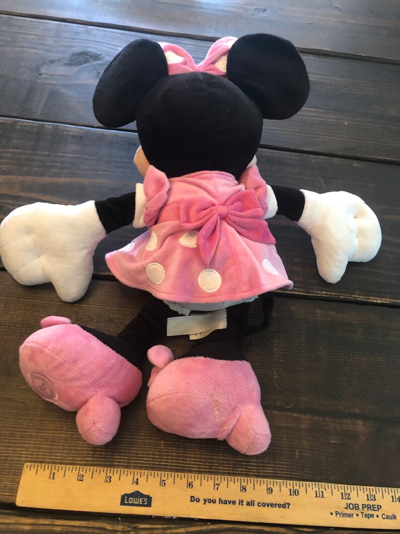 May include: A pink and black plush toy of Minnie Mouse wearing a pink polka dot dress with a pink bow. The toy is sitting on a wooden surface.