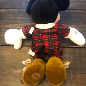 May include: A plush Mickey Mouse toy wearing a red and green plaid shirt and brown shoes. The toy is lying on its back with its arms outstretched.
